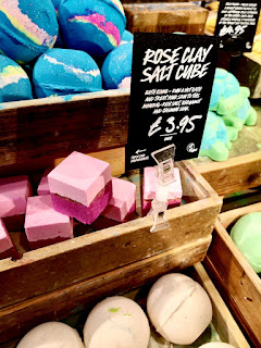 A group of light rose pink square bath bombs in a square light brown wooden box with a black rectangular label saying rose clay salt cube in white font on a bright background 