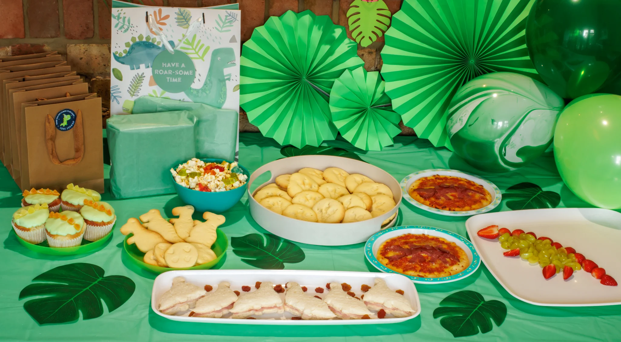 a table with dinosaur themed food, presents, balloons, green tablecloth and leaves