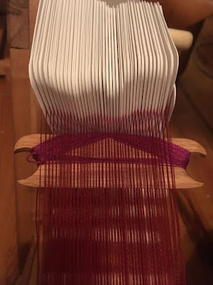 A shot from above of neatly arranged white tablet-weaving cards with spaced bands of extremely fine deep garnet thread emerging from between them, and a small, rectangular wooden shuttle with half-circles cut from each short end and wound with thicker magenta thread.