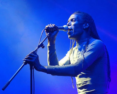 The Beat's Ranking Roger singing on stage as he holds the microphone and stand.