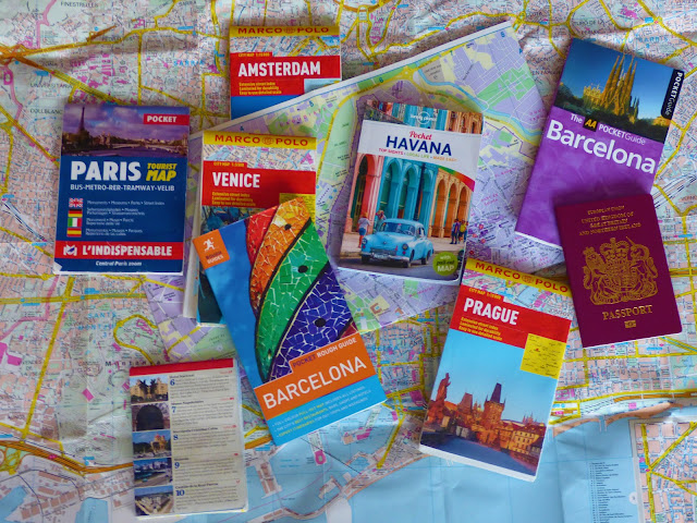 A flatlay of a map, plus travel maps and guidebooks for Paris, Barcelona, Prague, Marrakech, Havana and Venice 