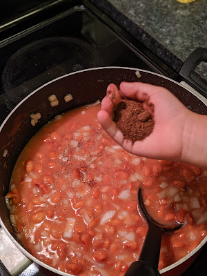 Image of girl adding handful of chili powder to the pot of chili on stove