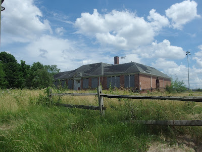 A boarded-up brick schoolbuilding sits in a field of overgrown grass and weeds. A falling apart wooden fence is in the foreground.