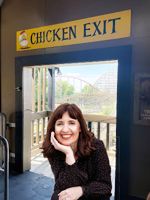 A white woman with dark hair in a dark, flower-print shirt sits in a doorway with a rollercoaster in the background. Above the doorway is a sign that says "Chicken Exit."
