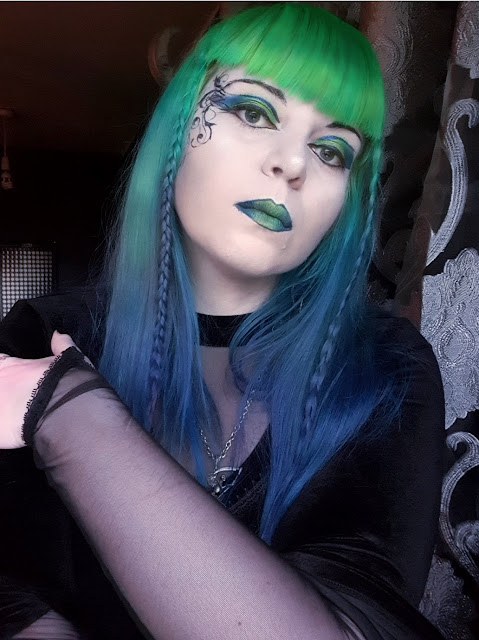 A woman with neon-green to royal blue gradient hair, neon green at the top, fading through emerald green and aqua to the deep blues of the ocean. She has pale skin, and dramatic make-up. She is wearing eyeshadow in neon green, emerald green and bright blue, with metalic violet and black eyeliner in a 'winged' style, and black mascara. She has vines in a swirling design drawn on the right side of her face. She is wearing green metallic lipstick outlined in metallic violet. She is wearing a high-necked velvet top with a v-neck style mesh insert, and with one arm across her body she is showing that the sleeves of the top are mesh, terminating in a point at her hand. She is wearing a pewter pendant of a winged Grim Reaper from Alchemy Gothic. Her hair is long, with a fringe and a thin braid either side of her face. She is in a room with dark purple walls.