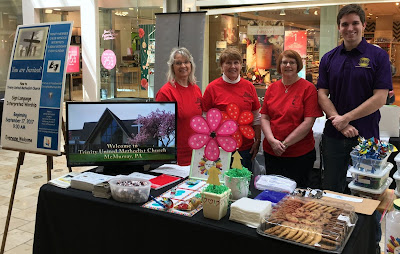 A group stands around a table in a shopping mall, there is a poster welcoming anyone to the church with the sign for ASL; they have cookies!