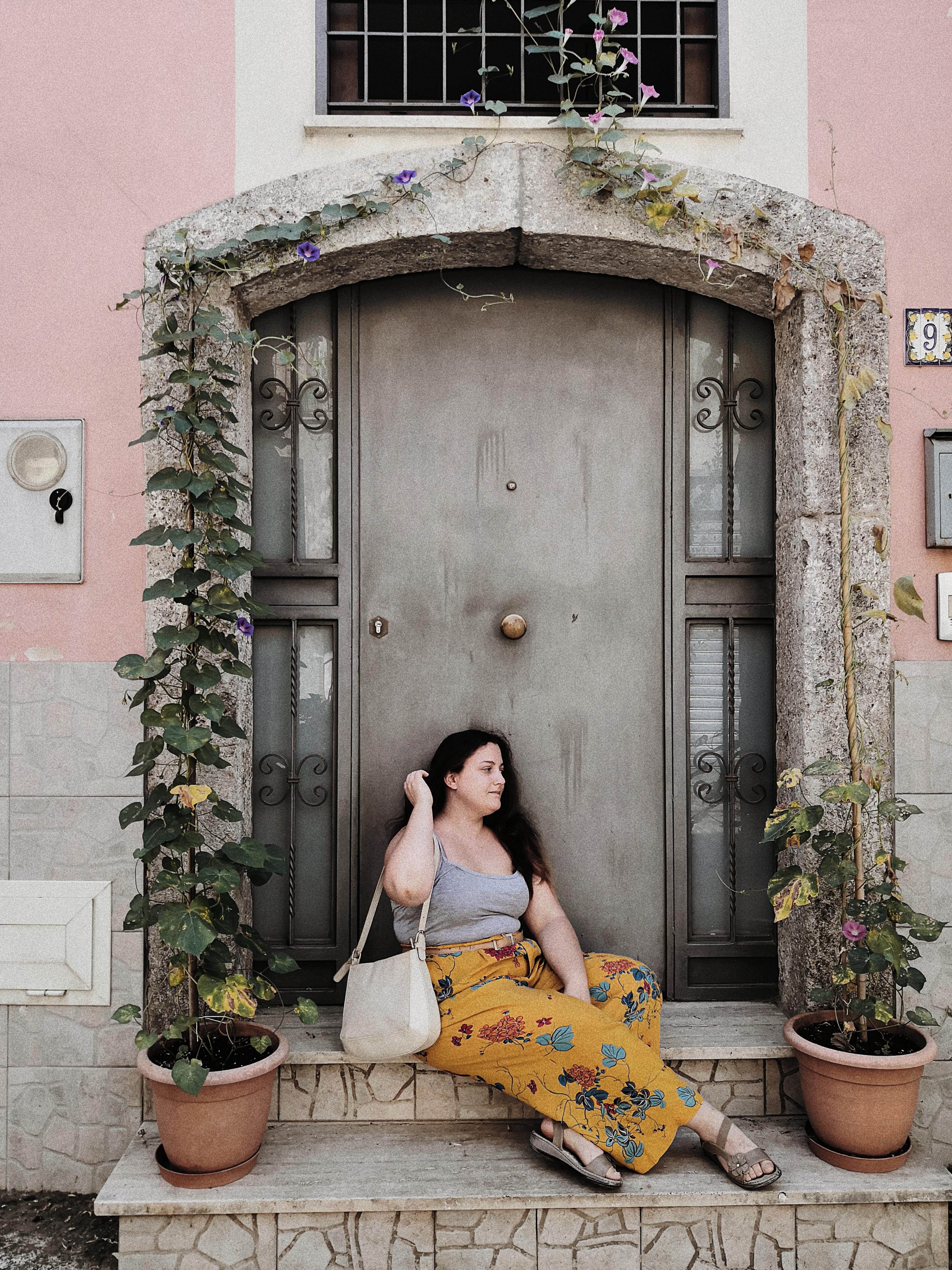 Ilaria è una ragazza di 32 anni, è seduta sulle scale davanti alla porta di casa sua. Indossa un paio di pantaloni floreali color senape e un top grigio. Ai piedi sandali bassi beige e una borsa a spalla beige chiaro. I capelli sono sciolti. Ai lati della porta di casa ha due vasi con delle ipomee che si arrampicano incorniciando la porta