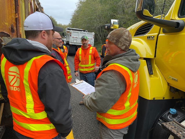 A hdnaful of people wearing safety orange reflective vests stand in a circle. There are large snowplows on either side of them. One person is talking and holding a clipboard.