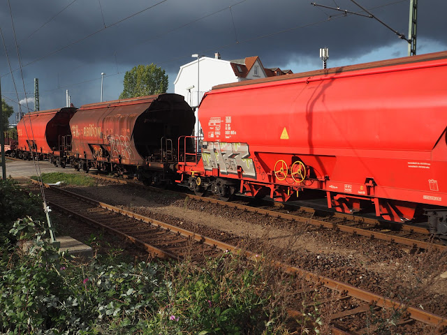 Deutlicher Kontrast: Leuchtend roter Waggon vorn, dahinter eher verrostet braun, dann wieder leuchtend rot von der Sonne angestrahlt. Darüber dunkle Regenwolken.