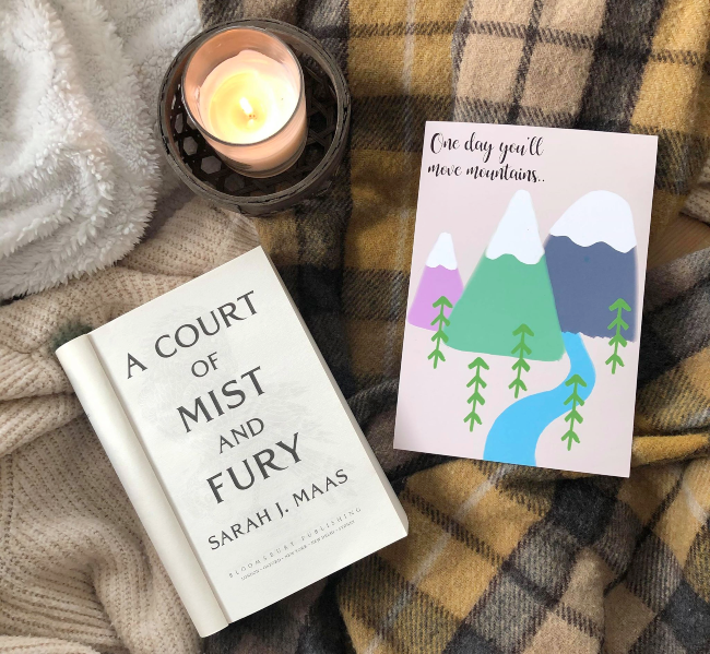 Title page of 'A COurt of Mist and Fury' next to a candle and art print of a mountain