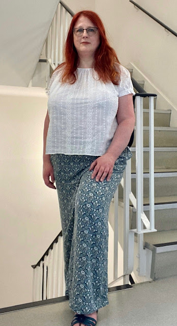 A redhaired white woman standing in an office stairwell. She is wearing a white embroidered top and print trousers. The top has short, slightly puffed sleeves and a gathered neckline, then falls straight, the gathered fabric smoothing out over the bust to show the delicate vertical stripes of the embroidery..