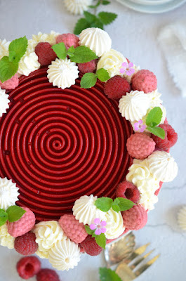 Himbeer-Dirndlmousse-Torte mit Brennnessel-Biskuit von oben fotografiert – zu sehen ist die tiefrote Himbeerspirale und rundherum Schlagoberstupfen, frische Himbeeren und Himbeeren aus Dirndlmousse, Baiser, Blätter und Blüten.