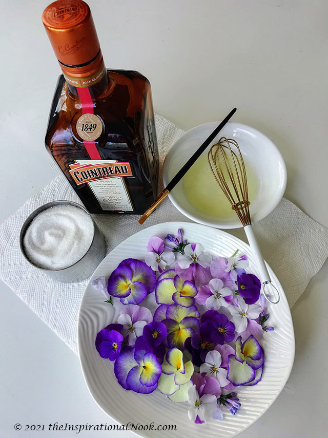 A bowls purple and yellow pansies, a whisk, a bottle of cointreau, sugar and egg whites to make candied pansies