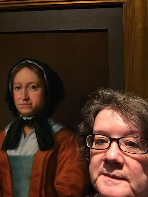 Selfie of me, a middle-aged white woman with brown and gray hair and glasses, and portrait of Hannah Penn, a white 17th-century woman wearing a dark head covering, white shift and orange gown. Photo shows definite similarities between the two of us, especially the laugh lines around our mouths.