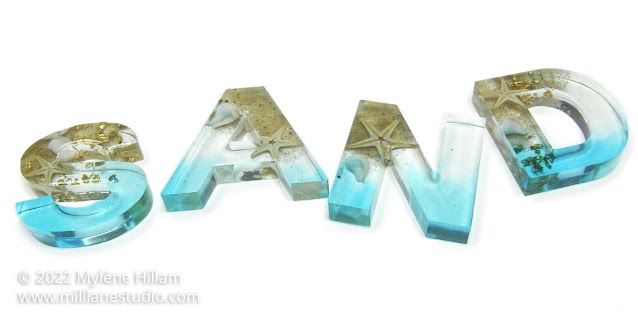 Resin letters spelling SAND filled with sand, starfish and goldleaf with a transparent blue background