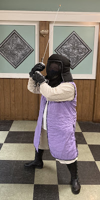 A person holding a longsword above their right shoulder. They are wearing a black fencing mask/hood, black gloves, black boots, and a white/lavender ensemble of a jacket, tunic, vest, and shorts.