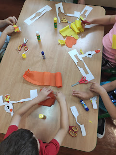 Na zdjęciu widzimy rączki dzieci, które na stoliku w kolorze drewna wykonują kolorowe prace plastyczne obrazujące uśmiechnięte duszki.