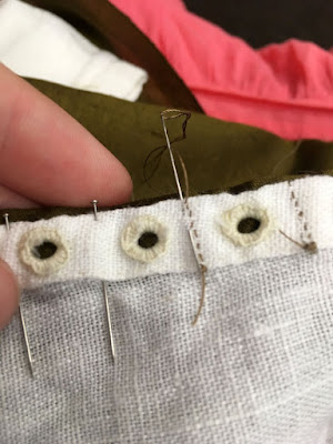 A close-up of neat white eyelets on a double-folded hem in white linen, with two lines of tiny tan stitches holding down the fabric between the right pairs of eyelets, and pins holding the fabric between the left pairs of eyelets. A needle in inserted from the edge of the fabric toward the inner fold of the hem.