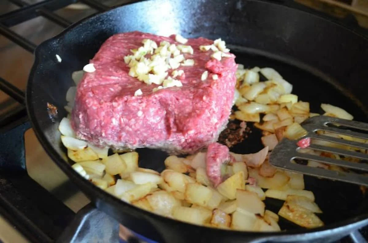 Sautéed onions in a cast iron skillet with a uncooked ground beef with minced garlic on top.