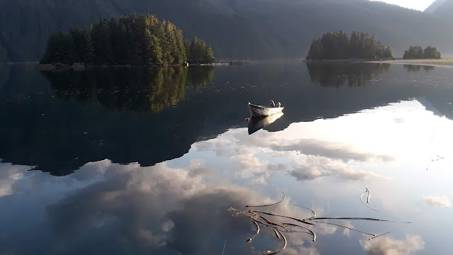 view of skiff in still water, islands, mountains, and reflection of clouds in water