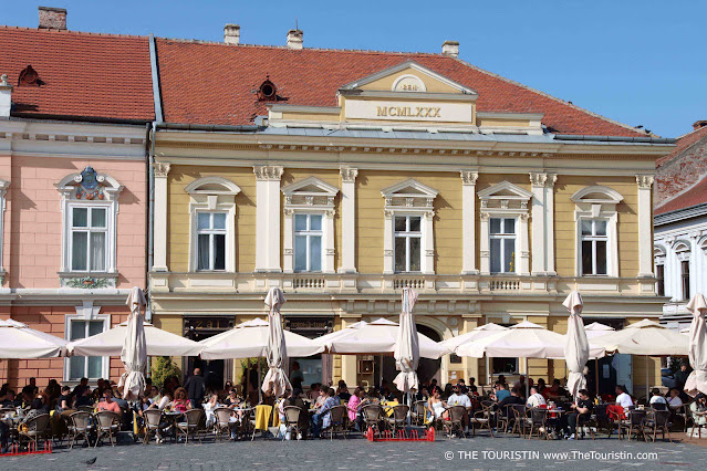 Cafes with white sun umbrellas where every single table with bistro chairs is occupied by hundreds of guests dressed in summer clothes in front of a row of yellow and salmon pastel-coloured two-storey period houses.