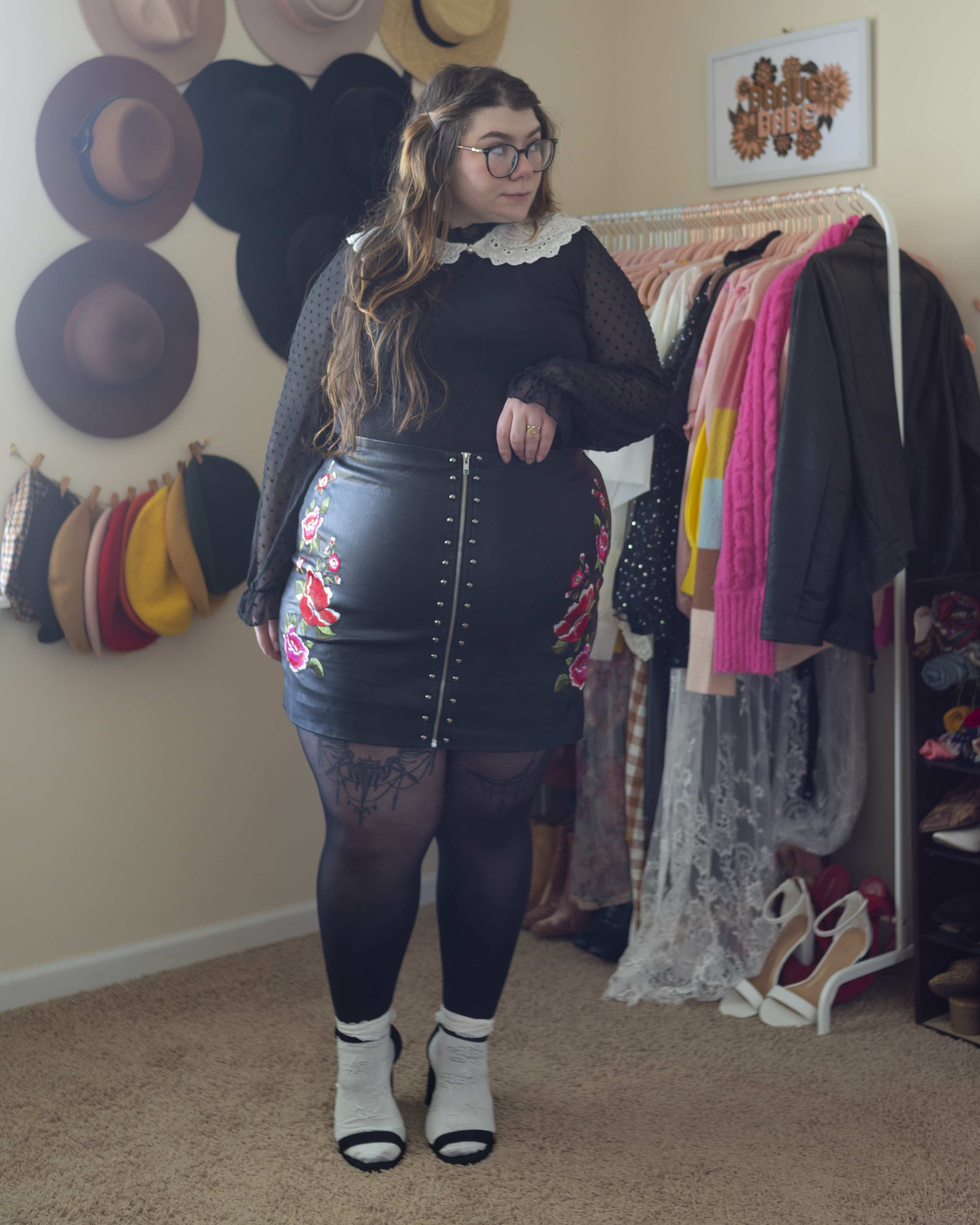 An outfit consisting of a white lace scalloped peter pan collar layered over a black long sleeve bodysuit with sheer black sleeves with polka dots tucked unto a black faux leather zip up mini skirt with red, hot pink, white and green embroidered flowers on the hips, black sheer tights, white lace ruffle socks and black ankle strap heels.