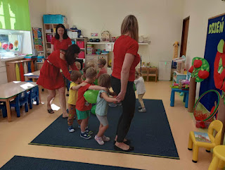 Sala przedszkolna. Przedstawia panią bibliotekarka i panie przedszkolanki z dziećmi, które stoją jedno za drugim między nimi są zielone balony.