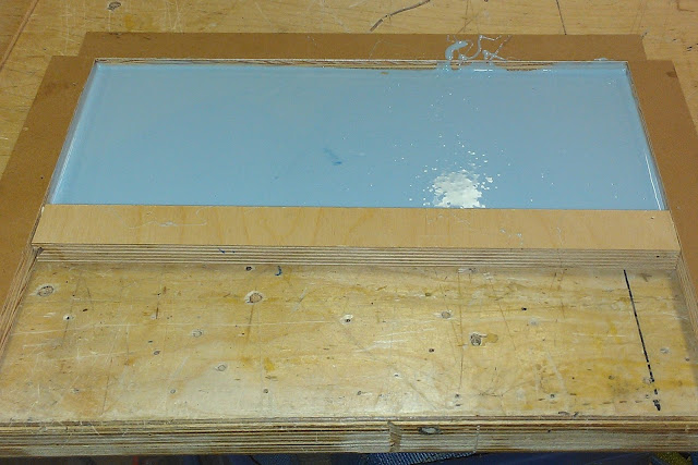 A rectangular box of about 5" by 9" sits on a wooden table.  In the box is the blue mold making material.  It is over a piece of plastic with the desired Braille for the exhibit text and will create the mold for the cast of the exhibit text that will go on display.