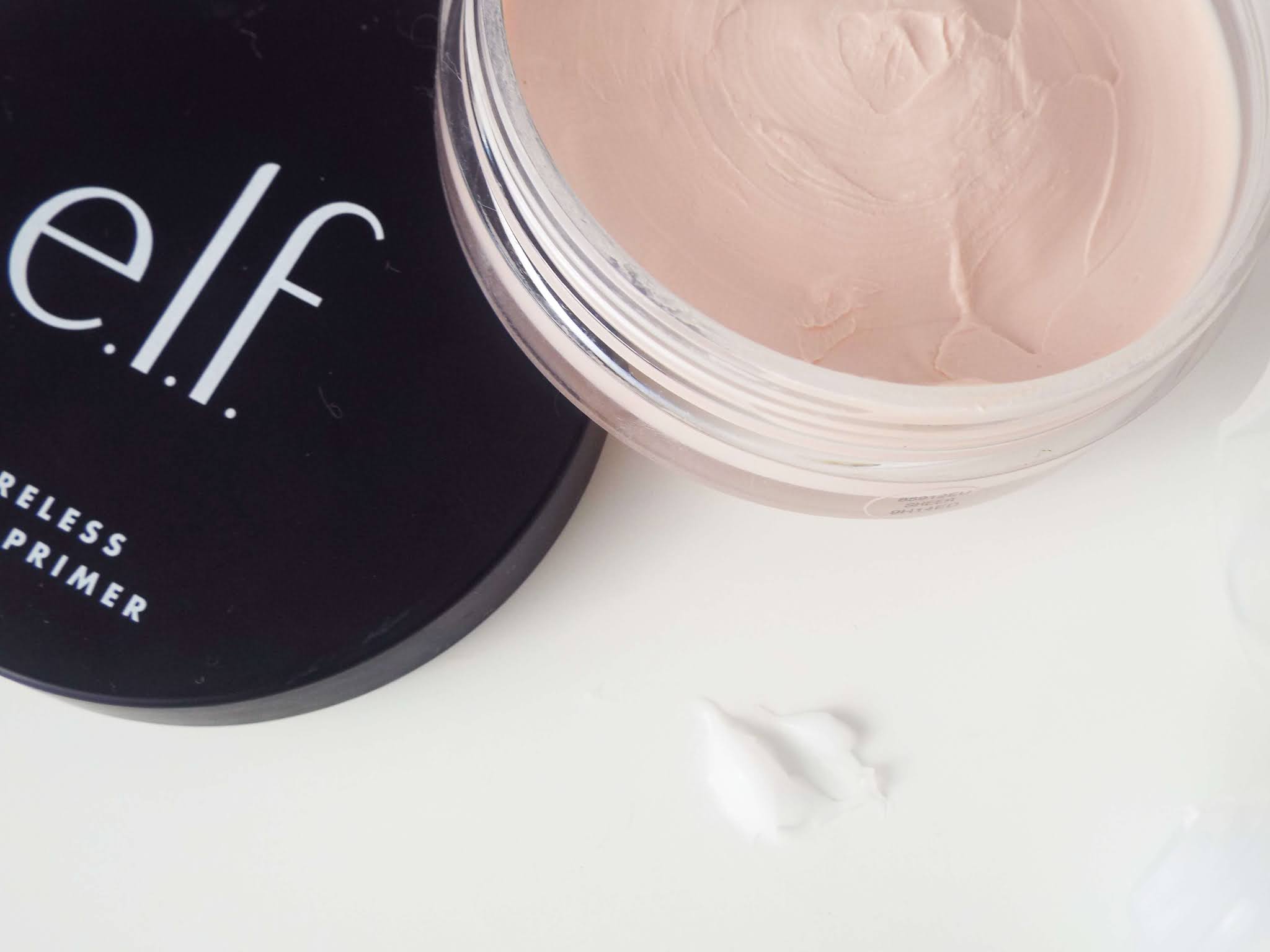 Ariel shot of the open Elf Poreless Putty Primer, with lid resting on side of pot and blob of The Ordinary High-Adherence Primer adjacent, comparing the difference in the thin, light pink Elf Poreless Putty Primer and thicker, white silicone The Ordinary Primer.