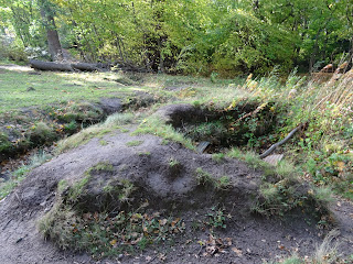 A photo of a muddy looking mound with grass growing out of a hollow in it.  This is one of the training trenches at Dreghorn Woods.  Photograph by Kevin Nosferatu for the Skulferatu Project.