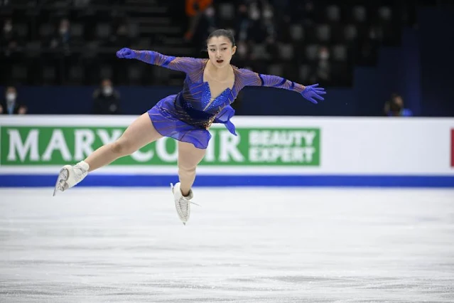 Sakamoto voa com seus patins sobre o gelo. Ela veste um vestido azul royal