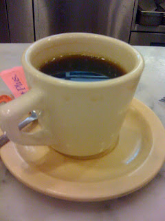 A still photograph of a full cup of black coffee (with a torn sweet and low on the saucer)