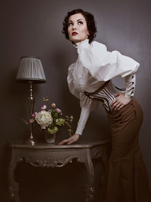 A woman clad in Neo-Vicotrian garb leans against a beautiful end table against a grey backdrop.