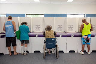 image shot from behind showing 4 men, one man a wheelchair user, voting at a polling place.