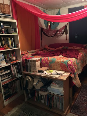 A room with a tall bed made up in fuchsia sheets and a bright pink, yellow, and turquoise print quilt in the corner, with a tall white bookshelf and short bronw bookshelf pressed against the foot of the bed and swags of pink, yellow, purple, and turquoise fabrics hanging from the windows and walls. The bookshelves are crammed with books and tchotchkes, and two Oriental rugs cover the wood floor at the foot and side of the bed.