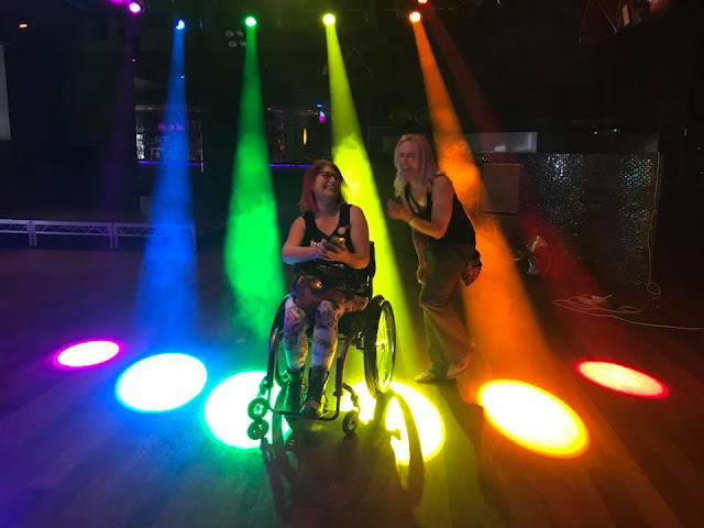 Two women are laughing.  One is a wheelchair user.  They are standing in puddles of different coloured light in the colours of the rainbow, caused by spotlights. 