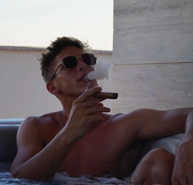 Shirtless dude in his 27 in a hot tub wearing sunglasses and smoking cigar