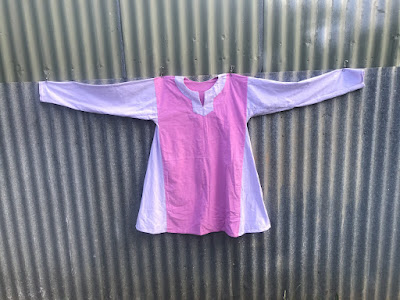 A tunic dress is pegged with its arms outspread on a corrugated iron fence. It has a keyhole neckline with a decorative facing on the outside. The body is a deep pink rectangle with triangular gores made of lavender fabric. The sleeves and facing are made of the same lavender fabric. End ID.