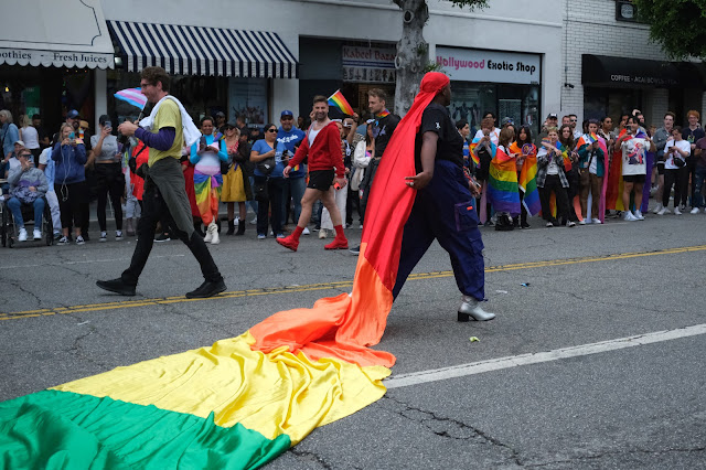 A man with a long fabric headpiece walks proudly in the pride parade.  Only red, orange, yellow, and green are visible in the photo.