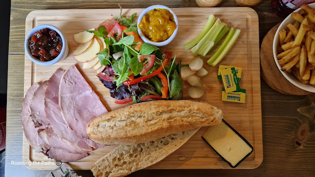 Lanscape shot of a Ploughmans Lunch from the Woolpack at Sopley. 4 slices of ham, a quarter of a small cheddar wheel, salad, pickle and picaclilli, salad, Pickled onions, warm half baguette on a wooded platter.