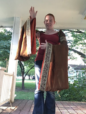 A yong white woman standing on a red porch with a white roof and plants in the background. Her red hair is pulled back, and she's wearing a dark red tank top and jeans. Her left arm is crooked in front of her and her right arm lifted, obscured by the long pendant sleeve in green-gold silk with peach lining and gold-floral-embroidered bands at the hems and upper arms.