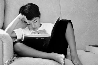 child with short dark hair sits on couch reading a book, with legs tucked up