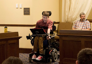 Justin presenting at Capitol using wheelchair and communication device, dad sitting in background