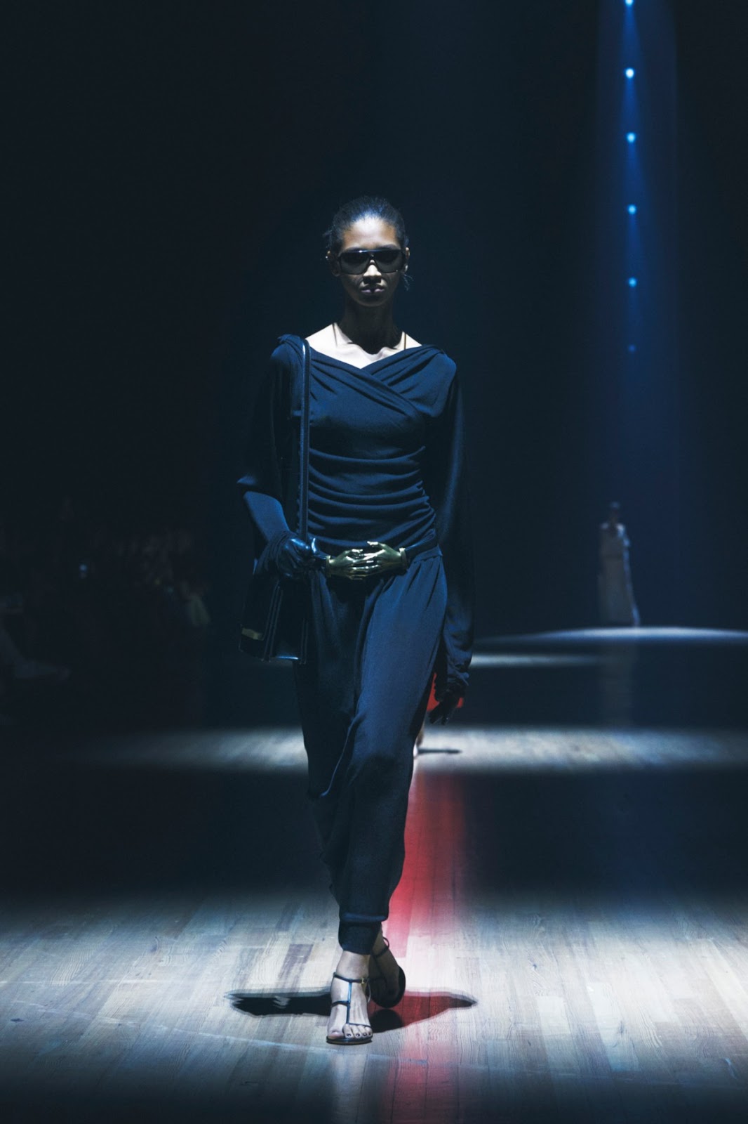 Woman with black hair in messy bun wearing black aviator sunglasses draping black maxidress with a black leather belt with gold clasped hands buckle, black gloves, black strappy high-heel sandals and a black leather shoulder bag walking on wooden floorboards under a spotlight