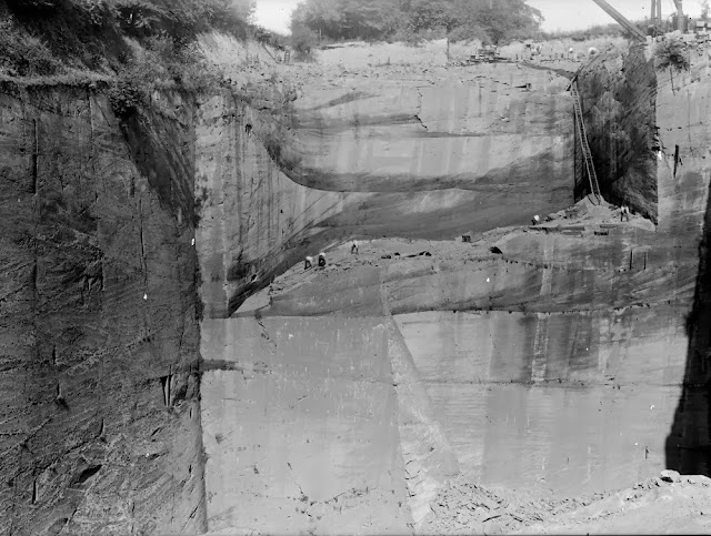 Ballochmyle Quarry, Mauchline. Ayrshire. New Red Sandstone (Permian) red desert sandstones showing large-scale aeolian (wind-blown) dune-bedding. The huge size of the quarry is indicated by the scale of the quarrymen – the quarry is often over 64 m deep. Access to the working face by ladder is clearly seen. Health & safety was not what is today! Large blocks are lifted by crane powered by a vertical steam boiler. The sandstone consists of well-sorted, mostly rounded and frosted sand grains. They are red in colour due to a coating of haematite, an iron oxide.