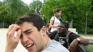 Foreground: Young man with short black hair and a grey t-shirt scratching is forehead with his eyes scrunched and mouth slightly open. Behind the confused looking man is another man in a black shirt looking off into the distance with a furrowed brow. Behind them is a woman in a white shirt with a dark grey scarf looking to the right. Behind them all is a lush green forest.