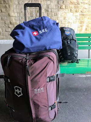 Suitcase with a blue tote bag on top. A black backpack sits on a bench behind the other two bags