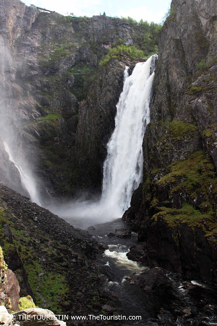 A giant waterfall crashes into a fast running mountain river that runs through a craggy mountain scape.