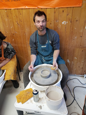 Image shows Max in an apron seated at a pottery wheel. In front of him is a bowl-shaped clay structure positioned in the center of the wheel.