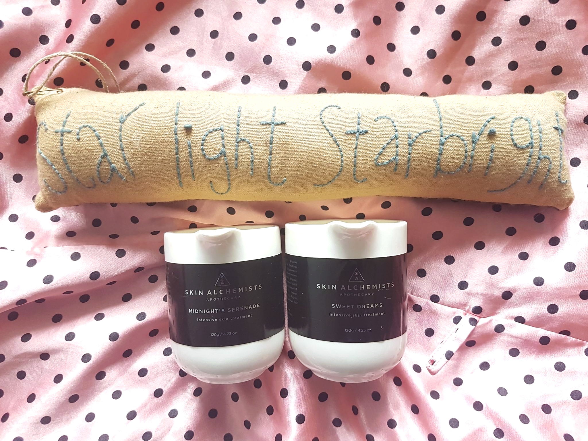 Skin Alchemists Intensive Skin Treatment Massage Candle on a pink silky robe with black polka dots with a long thin brown cushion that says star light star bright
